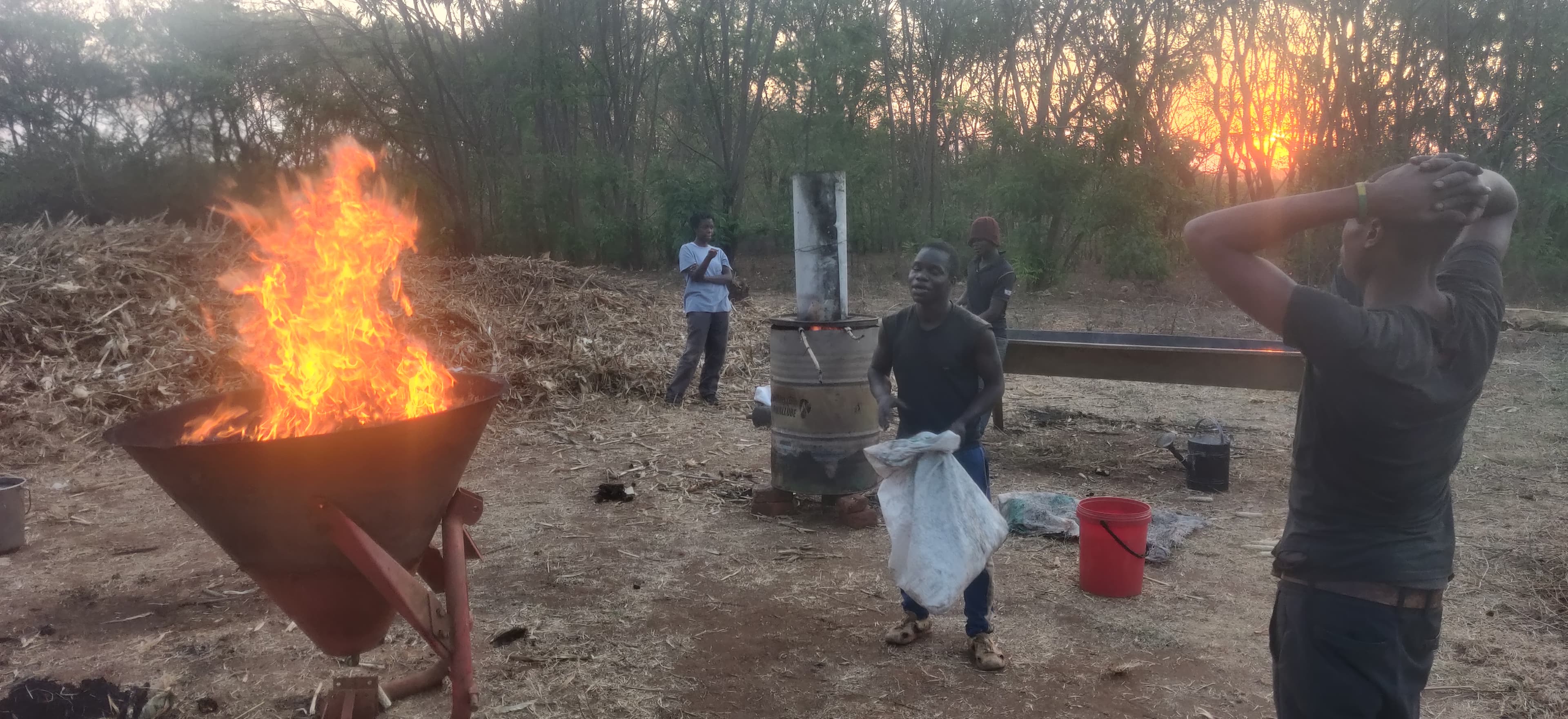 Hommes travaillant près d'un grand feu et d'un baril métallique au coucher du soleil.